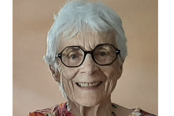 Elderly woman smiling, wearing glasses and a colorful top against a neutral background.